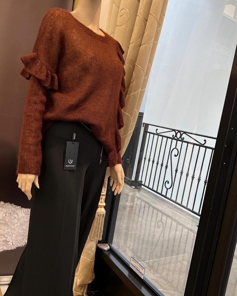 Puede incluir: Un su&eacute;ter de color &oacute;xido con mangas con volantes. El su&eacute;ter est&aacute; hecho de un material suave y peludo. El su&eacute;ter se est&aacute; exhibiendo en un maniqu&iacute; en una tienda.
