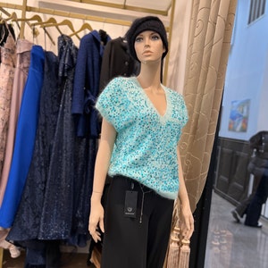 K&ouml;nnte beinhalten: Eine Schaufensterpuppe tr&auml;gt einen hellblauen, paillettenbesetzten Pullover mit V-Ausschnitt und kurzen &Auml;rmeln, schwarze Hosen und eine schwarze Baskenm&uuml;tze. Die Puppe steht vor einem Kleiderst&auml;nder.