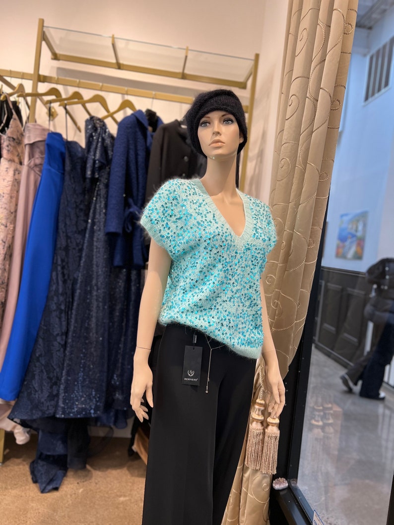 K&ouml;nnte beinhalten: Eine Schaufensterpuppe tr&auml;gt einen hellblauen, paillettenbesetzten Pullover mit V-Ausschnitt und kurzen &Auml;rmeln, schwarze Hosen und eine schwarze Baskenm&uuml;tze. Die Puppe steht vor einem Kleiderst&auml;nder.