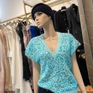 K&ouml;nnte beinhalten: Ein hellblauer, paillettenbesetzter, &auml;rmelloser Pullover mit V-Ausschnitt. Der Pullover ist aus einem weichen, flauschigen Material gefertigt und hat eine glitzernde Oberfl&auml;che.