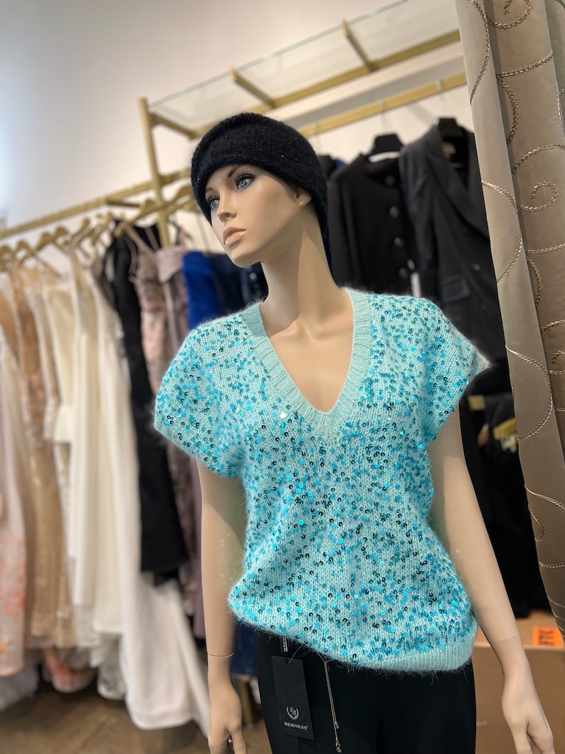 K&ouml;nnte beinhalten: Ein hellblauer, paillettenbesetzter, &auml;rmelloser Pullover mit V-Ausschnitt. Der Pullover ist aus einem weichen, flauschigen Material gefertigt und hat eine glitzernde Oberfl&auml;che.