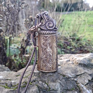 May include: A detailed, antique-style metal pendant with a cylindrical body and a decorative top featuring an animal head design. The pendant has intricate Celtic knot patterns and a brown leather strap. The overall color scheme is metallic silver and bronze.