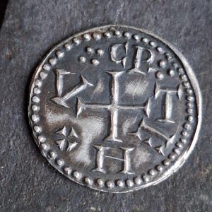 May include: A silver coin with a cross design and the letters "CPT" and "H". The coin has a beaded border and is likely an antique or replica. The coin is circular and has a worn appearance.