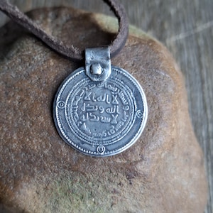 May include: A silver pendant with Arabic script, attached to a brown leather cord. The circular pendant has a detailed design with a central inscription and an outer border. The pendant is displayed on a textured, brown surface.