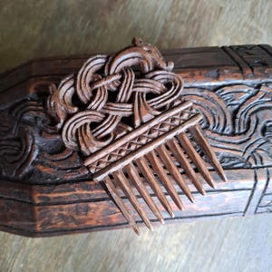 May include: A dark brown wooden hair comb featuring an intricate carved knotwork design at the top, which incorporates two stylised animal heads facing outward. The comb has a horizontal band with a geometric pattern above eight pointed teeth.