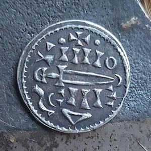 Könnte beinhalten: Eine silberne Münze mit aufwendigen Gravuren, darunter ein Schwert, Kreuze und andere Symbole. Die Münze hat einen gezahnten Rand und scheint antik zu sein. Sie liegt auf einer dunklen, strukturierten Oberfläche.
