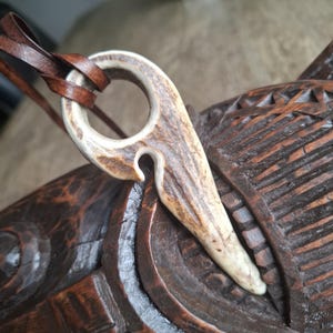 May include: A pendant crafted from a piece of natural antler with a textured, light brown and cream surface. The pendant features a circular top opening and a hook-like shape, suspended from a dark brown leather cord for use as a necklace.