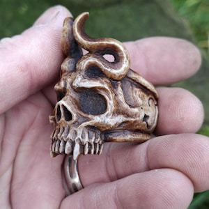 May include: A detailed, brown skull ring with curved horns. The ring features a detailed skull design with visible teeth and eye sockets. The ring is held in a hand, showcasing the intricate carving and the ring's overall design.