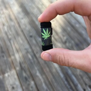May include: A small black and green tube with a marijuana leaf design. The tube is about the size of a finger.
