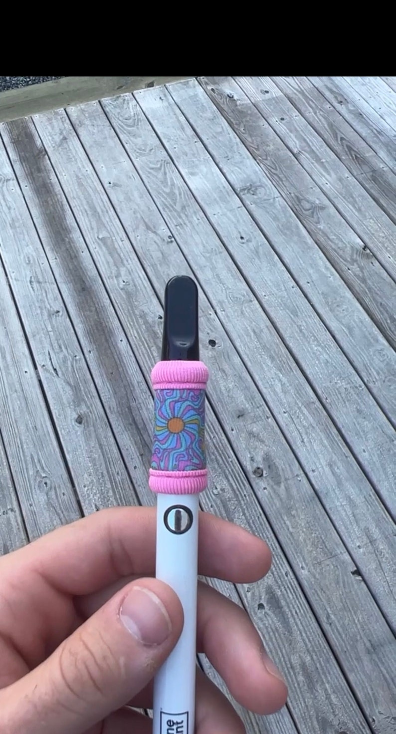 May include: A white vape pen with a pink and blue patterned sleeve. The sleeve has a flower design with a yellow center.