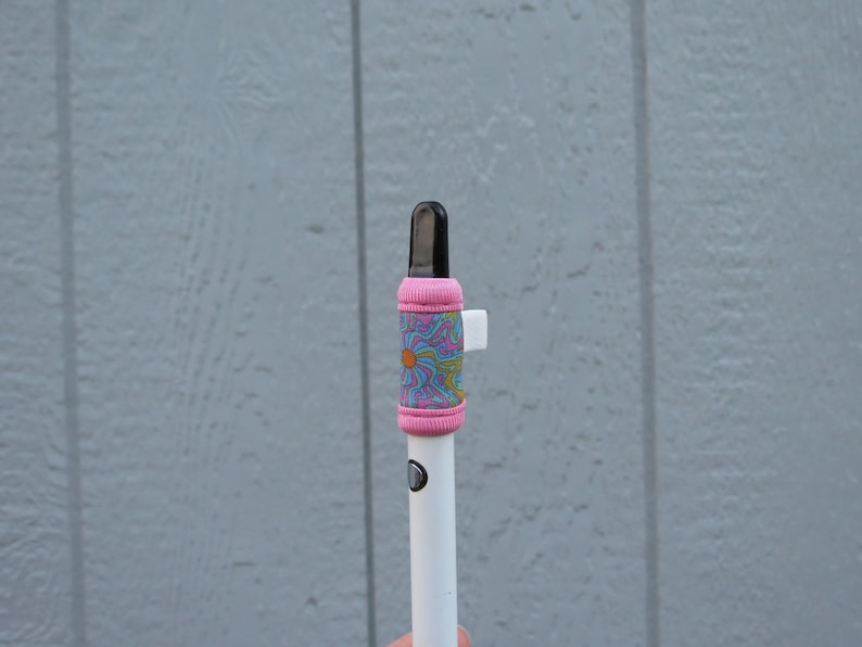 May include: A white vape pen with a pink and blue patterned sleeve. The sleeve has a white tab on the side.