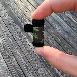 May include: A small black and green cylindrical container with a black elastic band. The container has a pattern of green marijuana leaves on a black background.