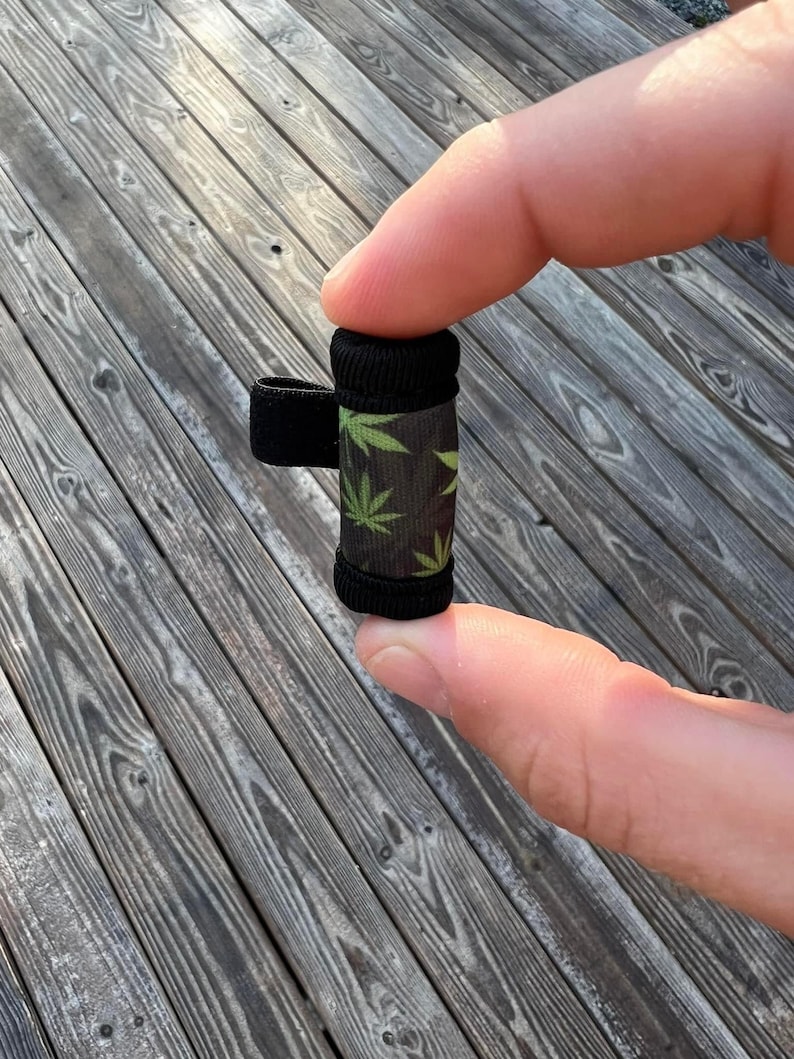 May include: A small black and green cylindrical container with a black elastic band. The container has a pattern of green marijuana leaves on a black background.