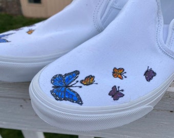 Könnte beinhalten: Weiße Slip-on-Canvas-Schuhe mit handbemalten Schmetterlingsmotiven. Die Schmetterlinge sind in Blautönen, Orange und Lila.