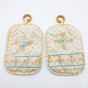 May include: Two quilted oven mitts with a floral pattern in pastel colors. Each mitt has a wooden ring for hanging and is trimmed with yellow binding. The design includes the text "Paul Revere".