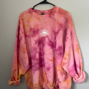 May include: A pink, orange, and purple tie-dye sweatshirt with a white sun graphic on the front.