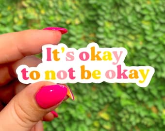 May include: A white sticker with the text "It's okay to not be okay" in pink, yellow, and orange letters. The sticker is being held by a hand with pink nail polish.