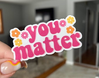 May include: Pink and white sticker with the text "you matter" in a retro font. The sticker is decorated with small orange and yellow flowers.