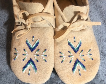 Könnte beinhalten: Ein Paar hellbraune Wildleder-Mokassins. Jeder Schuh hat ein Perlenmuster in Blau, Grün und Weiß. Die Mokassins haben eine weiche, runde Spitze und Lederschnürsenkel. Der Stil erinnert an indianisches Schuhwerk.