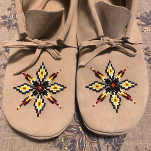 Könnte beinhalten: Ein Paar hellbraune Wildleder-Mokassins. Jeder Mokassin hat ein aufgesticktes Blumenmuster in Rot, Gelb, Schwarz und Weiß. Die Mokassins haben eine weiche, abgerundete Spitze und einen Kordelverschluss.
