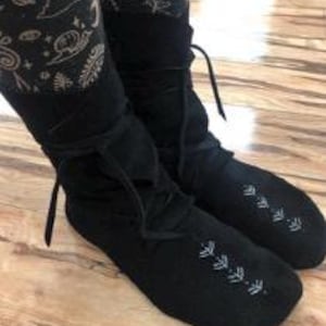 May include: A pair of black suede boots with a lace-up design. The boots have a soft, slouchy silhouette and feature decorative stitching along the toe. The boots are shown on a wooden surface.