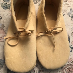 May include: A pair of light tan suede moccasin-style shoes. The shoes have a soft, textured appearance with a rounded toe and a low ankle. Leather laces are tied at the top of the foot. The shoes appear to be unworn.