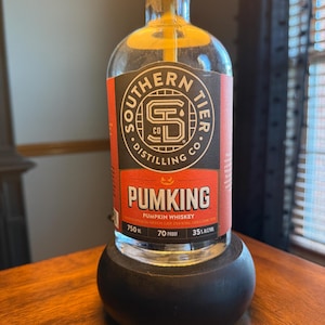 May include: A clear glass bottle of Southern Tier Distilling Co. Pumking Pumpkin Whiskey, with a black and orange label. The bottle is mounted on a dark wooden base, and the lamp is lit with a warm glow.