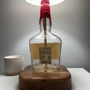 May include: A unique lamp made from a repurposed liquor bottle. The clear glass bottle has a red wax seal around the neck and sits on a wooden base. A beige lampshade sits above the bottle. The lamp is plugged in with a yellow cord.