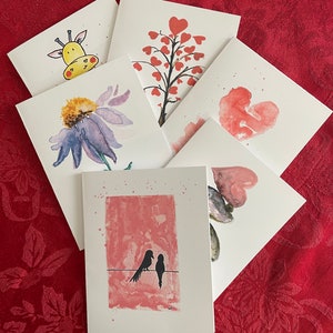 May include: Set of six blank greeting cards with watercolor illustrations. The cards feature a giraffe, a flower, a heart tree, a heart shape, two birds on a wire, and a heart shape with a fish.