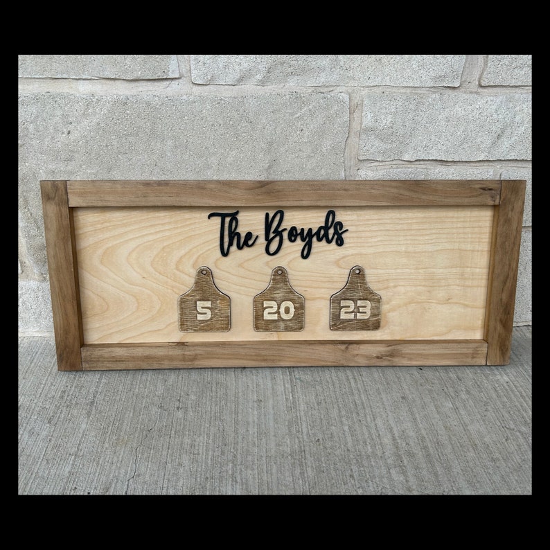 May include: A wooden sign with the text "The Boyds" in black cursive lettering. The sign has three wooden tags with the numbers 5, 20, and 23.