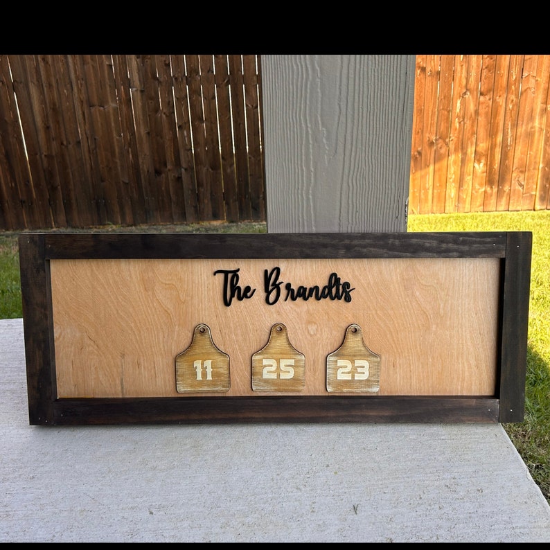 May include: A wooden sign with a dark brown frame and a light wood background. The sign says "The Brandts" in black cursive lettering. There are three wooden tags with white numbers on them: 11, 25, and 23.