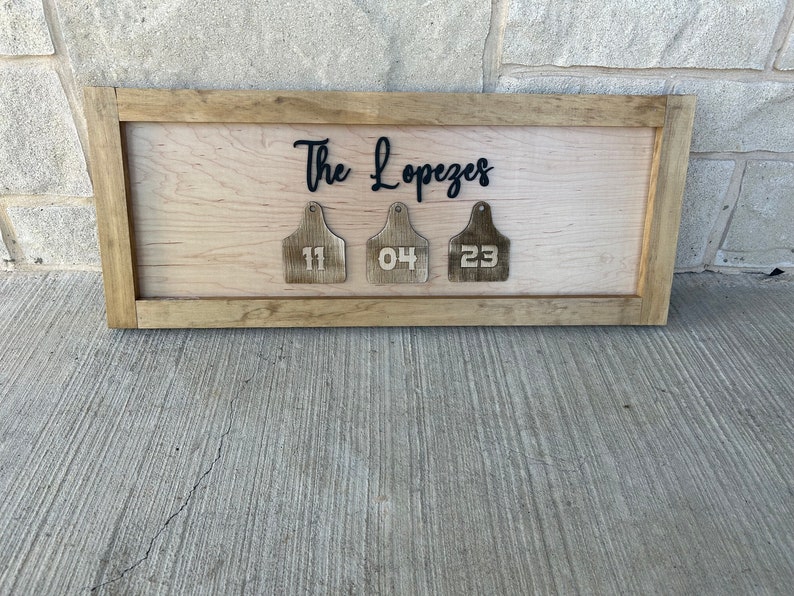 May include: A wooden sign with the text "The Lopezes" and three wooden tags with the numbers "11", "04", and "23" hanging from it.