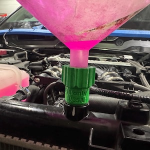 May include: A green and black radiator fill adapter with the text "Fill only V35A" is connected to a car's radiator. A pink funnel is inserted into the adapter, pouring a pink liquid into the engine. The car's engine bay is visible in the background.