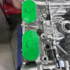 May include: Close-up of a metallic engine block with two bright green caps. The caps are oval-shaped and have the text "TOY/SUB", "ADC", and "FR24D" printed on them. The engine block has various holes and fittings.