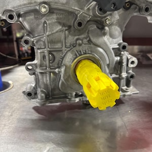 May include: A close-up of a yellow metal part with the text "2GR 1UR 3UR" on it. The part is attached to a silver engine block with the text "TOYOTA" visible.