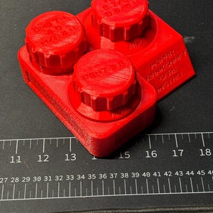 May include: A bright red 3D-printed automotive part with three circular knobs and a rectangular base. The part has text that reads "DMB TH M/REM", "HIGH 5.7/61 06/54", "PENT.3.6", "MOPAR TANKSHAFT SEAL INSTALL".