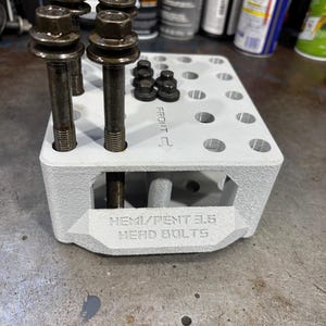 Puede incluir: Un organizador gris claro impreso en 3D que contiene varios pernos y tuercas de metal oscuro. El organizador tiene el texto "HEMI/PENT 3.6 HEAD BOLTS" y una flecha que indica el frente. Los pernos tienen cabezas hexagonales grandes.