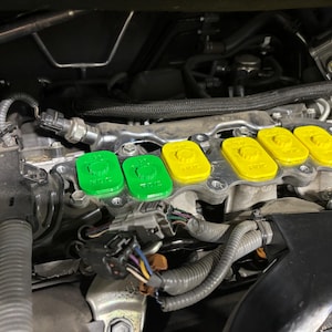 May include: An engine bay with green and yellow plastic caps installed. The caps are rectangular with a circular design on top. The engine components are visible in the background.