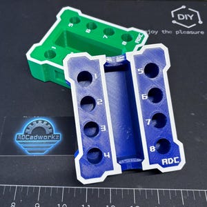 Pode incluir: Duas gabaritos impressos em 3D, um verde e outro azul, com detalhes brancos. Cada gabarito tem oito furos numerados. O gabarito azul tem o texto "ADC" e o gabarito verde tem o texto "5 6 7 8". O fundo é uma superfície preta com marcas de régua.