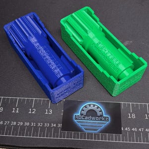 Pode incluir: Dois objetos de plástico impressos em 3D, um azul e outro verde, cada um em uma caixa retangular correspondente. O objeto azul tem o texto "Penbasba 518477818 sbar 3.G" impresso. Um cartão de visita com o logotipo "ADCadworkz" está em primeiro plano.