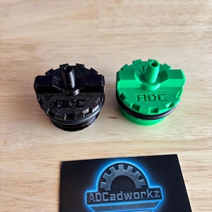 Puede incluir: Dos tapas impresas en 3D, una negra y otra verde, con un diseño texturizado y una boquilla central. La tapa verde tiene el texto "ADE". También hay una tarjeta de visita con el logotipo "ADCadworkz".