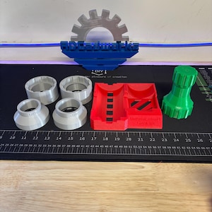 以下が含まれることがあります： さまざまな色の3Dプリントされたオブジェクトのコレクション。銀色と灰色のソケットのような部品、赤い長方形のホルダー、緑色の円筒形のアイテムが含まれています。オブジェクトは、定規が付いた黒い表面に配置されています。