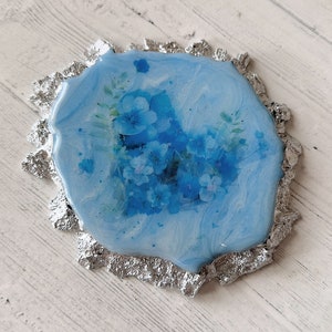 Pode incluir: Um descanso de copo de resina azul com uma borda de folha de prata e um design floral com flores azuis.