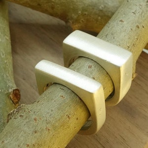 May include: Two silver square rings stacked on top of each other. The rings are on a light brown wooden surface.