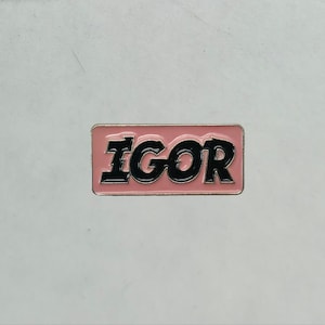 May include: A rectangular pin with the word "IGOR" in black, outlined in black, set against a pink background. The pin has a silver border and is set against a light gray background.
