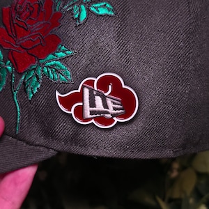 May include: A dark gray baseball cap with a red and white pin featuring a cloud design and a logo. The cap has a large embroidered red rose with green leaves. The pin is the focal point of the image.