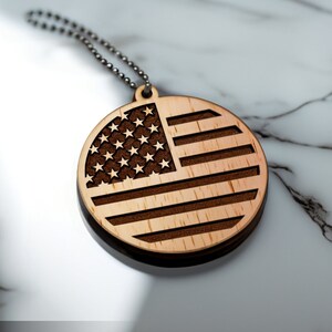 May include: A round wooden ornament featuring the American flag design. The flag is laser-cut into the wood, with stars and stripes visible. A metal ball chain is attached for hanging. The ornament is light brown with dark brown accents.