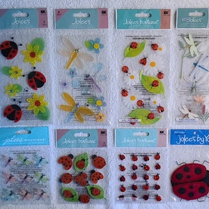 Könnte beinhalten: Ein Set mit 10 Blättern bunter, dekorativer Aufkleber mit Marienkäfern, Schmetterlingen und Blumen. Die Aufkleber sind in durchsichtigen Plastiktüten verpackt, auf denen der Markenname "Jolee's" und "Jolee's Boutique" steht.