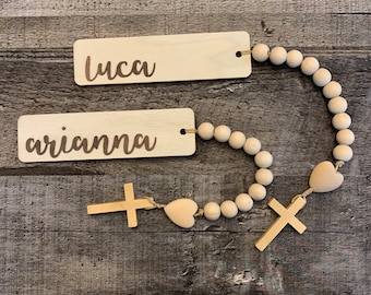 May include: Wooden bead garland with two wooden name tags, one with the name "luca" and the other with the name "arianna". The garland has wooden hearts and crosses.