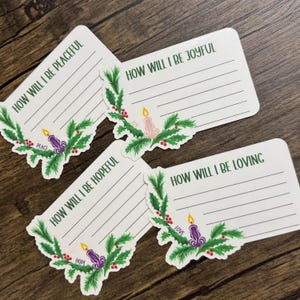 Peut inclure: Cartes rectangulaires blanches aux coins arrondis, avec le texte "HOW WILL I BE PEACEFUL", "HOW WILL I BE HOPEFUL", "HOW WILL I BE JOYFUL" et "HOW WILL I BE LOVING". Chaque carte a une bordure décorative de feuilles vertes, de baies rouges et d'une bougie.